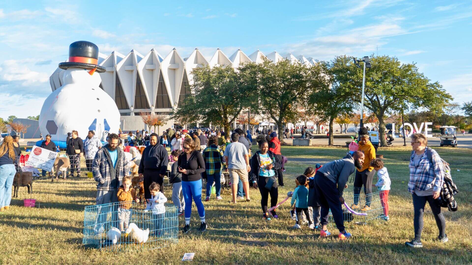 Families gather around petting zoo animals, some waiting in line for giant inflatable snowman bounce house, with the Hampton Coliseum in the background.