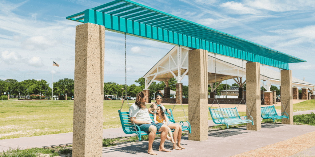 A family sits on turquoise swings under a shaded structure on a sunny day at Buckroe Beach & Park. The setting is relaxed, with a grassy field and pavilion in the background.