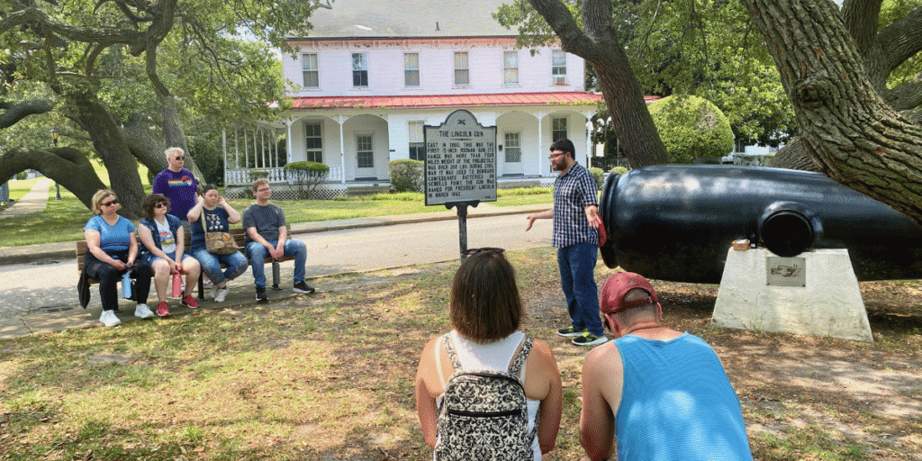 Under a large shady tree, a tour guide speaks to groups of families seated on benches next to historical marker and the Lincoln Gun at Fort Monroe.