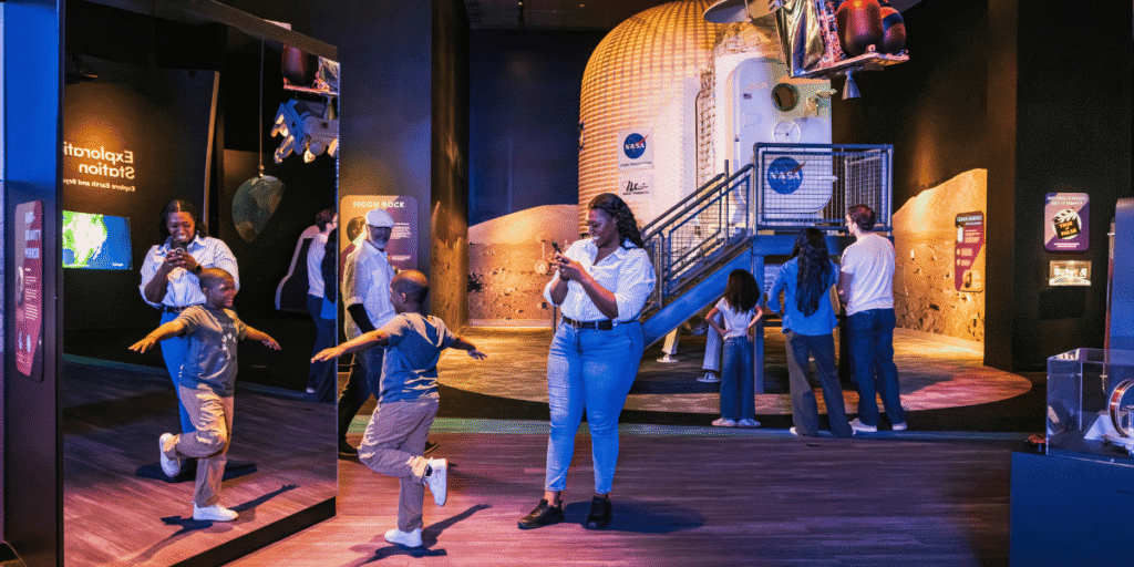 A mother takes pictures of her son playing in front of an interactive exhibit. In the background, another family looks at a life-size lunar landing pod.