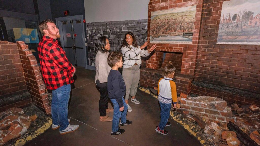 A family listens to a guide explaining historical paintings at Hampton History Museum in an exhibit that recreates the scene of burnt rubble from brick buildings. The scene appears educational and engaging.