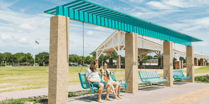 A family sits on turquoise swings under a shaded structure on a sunny day at Buckroe Beach & Park. The setting is relaxed, with a grassy field and pavilion in the background.