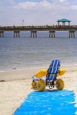Sand-ready WaterWheels Floating Beach Wheelchair at the end of a blue Mobimat Roll-out ADA Beach access pathway at Buckroe Beach in Hampton with the fishing pier in the background.