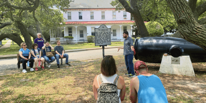Under a large shady tree, a tour guide speaks to groups of families seated on benches next to historical marker and the Lincoln Gun at Fort Monroe.