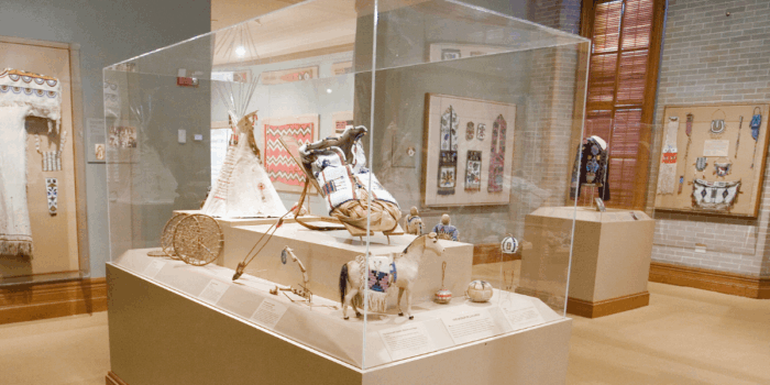 Museum exhibit showcasing Native American artifacts & recreations, including a cradleboard, hoop & pole game, horse, dolls, and other handmade toys, enclosed in a glass display case. In the background wearable artifacts like tunics, jewelry, & bags are displayed.