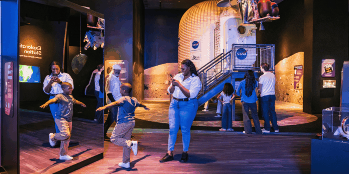 A mother takes pictures of her son playing in front of an interactive exhibit. In the background, another family looks at a life-size lunar landing pod.