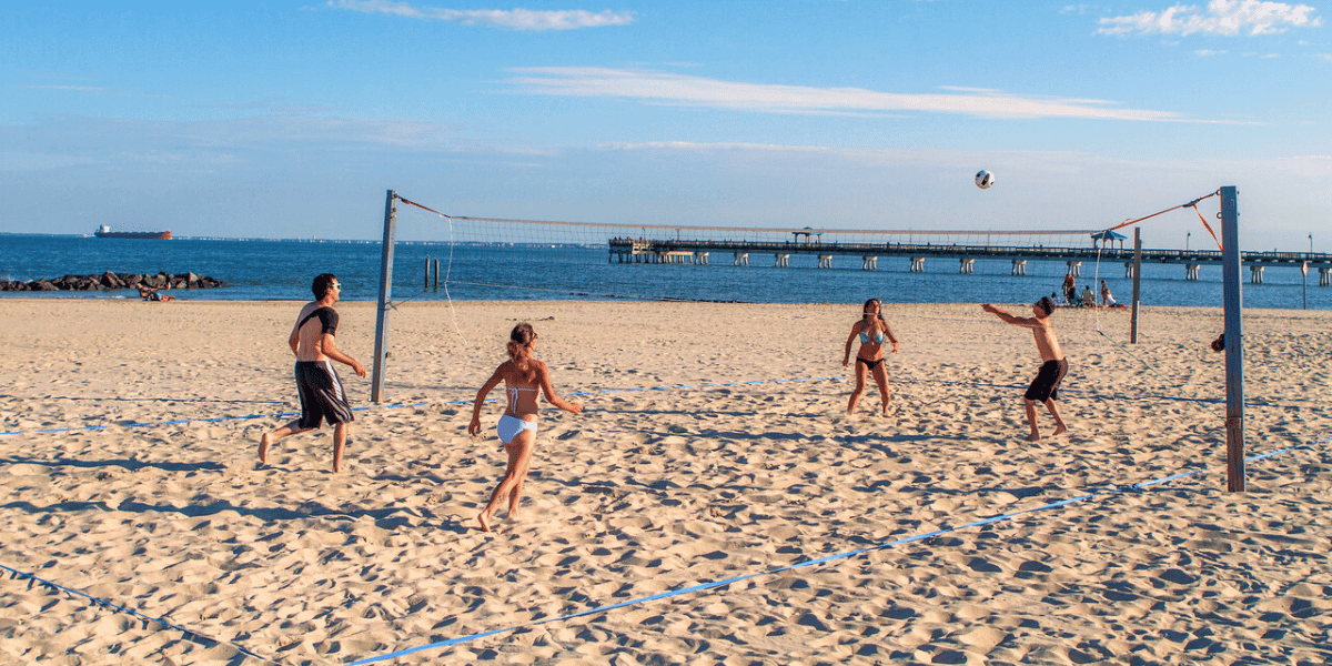 Young adults play sand volleyball at Buckroe Beach with fishing pier and shipping boat in background