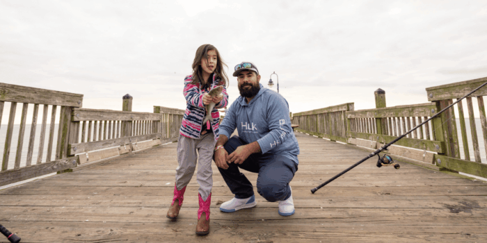 A young girl shows off the fish she just caught. She's standing next to her father on the wooden fishing pier with a fishing rod nearby.