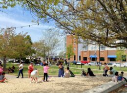 A lively park scene shows children playing and adults chatting under trees. A building and parked cars are visible in the background, evoking a relaxed, sunny day.