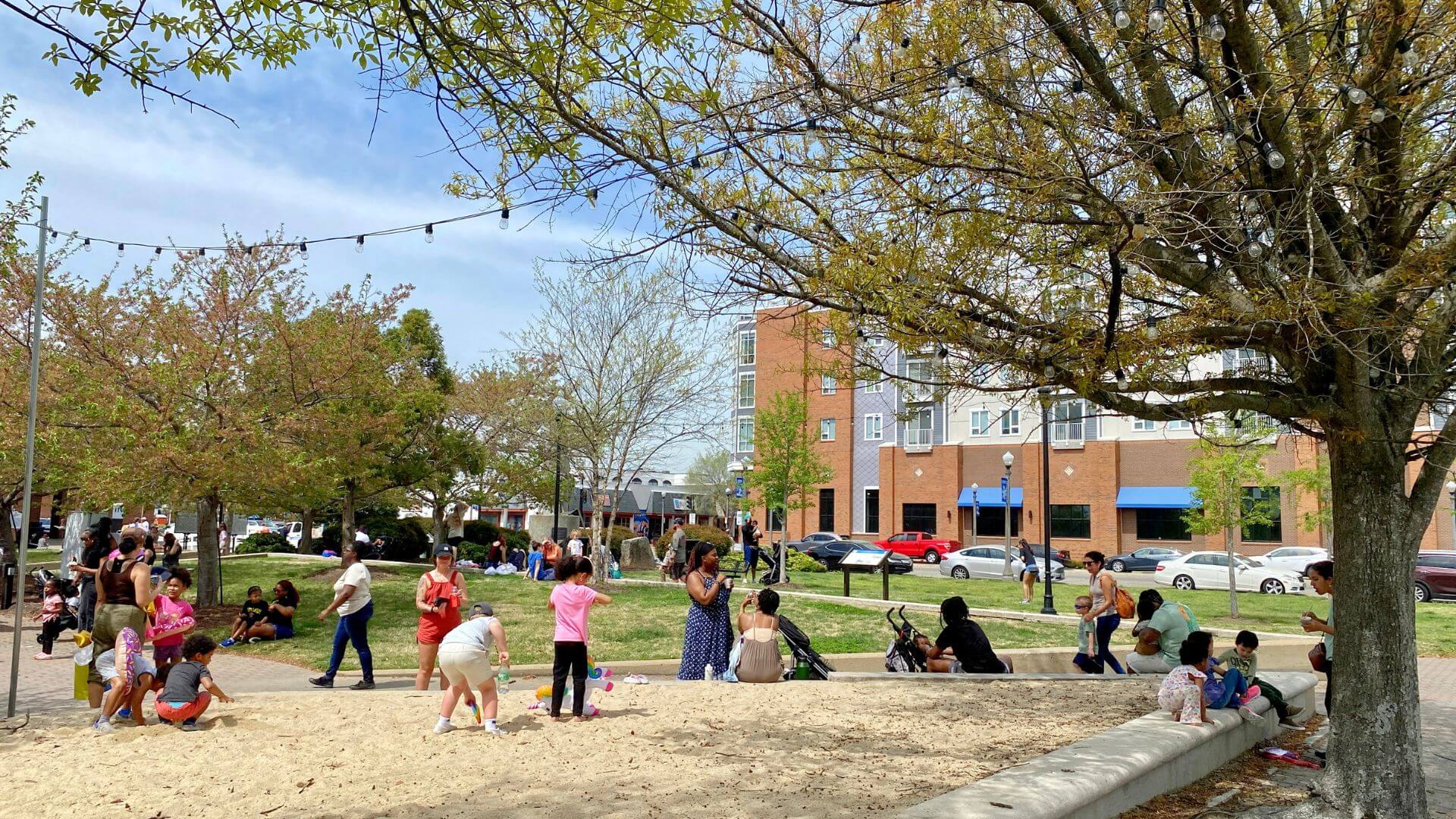 A lively park scene shows children playing and adults chatting under trees. A building and parked cars are visible in the background, evoking a relaxed, sunny day.