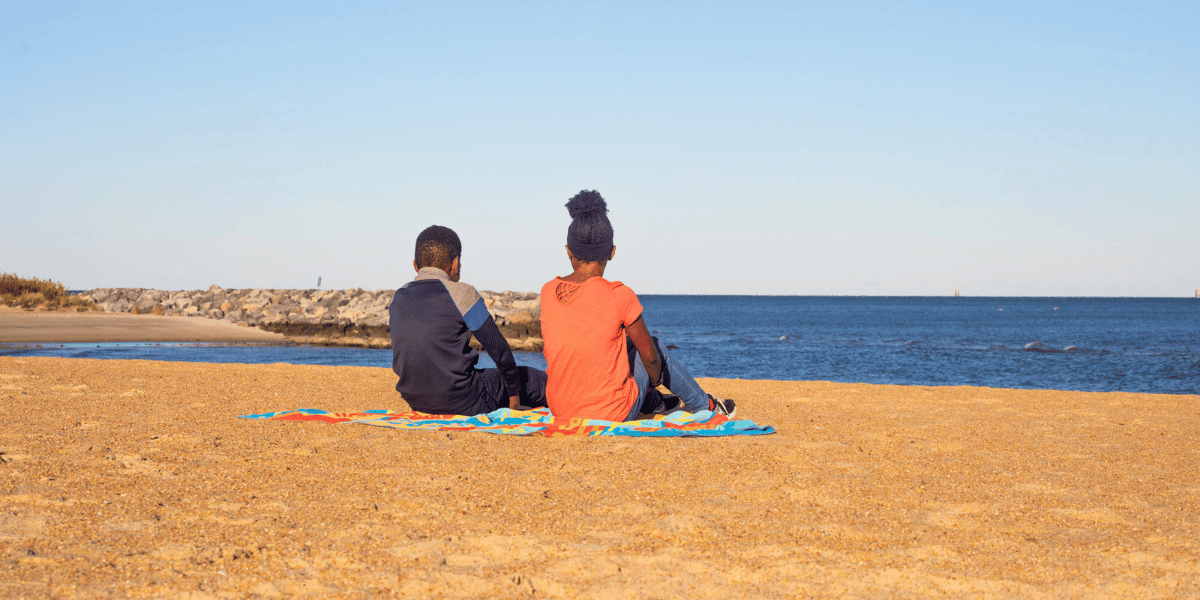 Two kids sit on a beach towel as they watch the water at Outlook Beach in Fort Monroe
