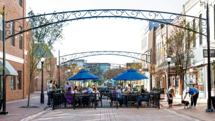People enjoy a sunny day at an outdoor streatery with blue umbrellas, surrounded by brick buildings and decorative arches, creating a lively, relaxed atmosphere.