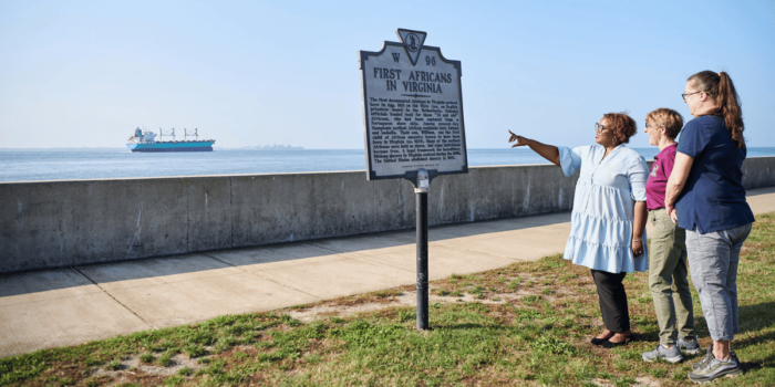 Three women read the First Africans in Virginia historical marker by the Seawall in Fort Monroe as a ship passes by in the distance 